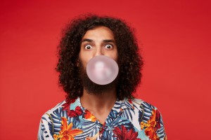 Astonished attractive young brunette curly man with beard wearing multi-colored flowered shirt while posing over red background, rounding eyes and inflating bubble with pink gum